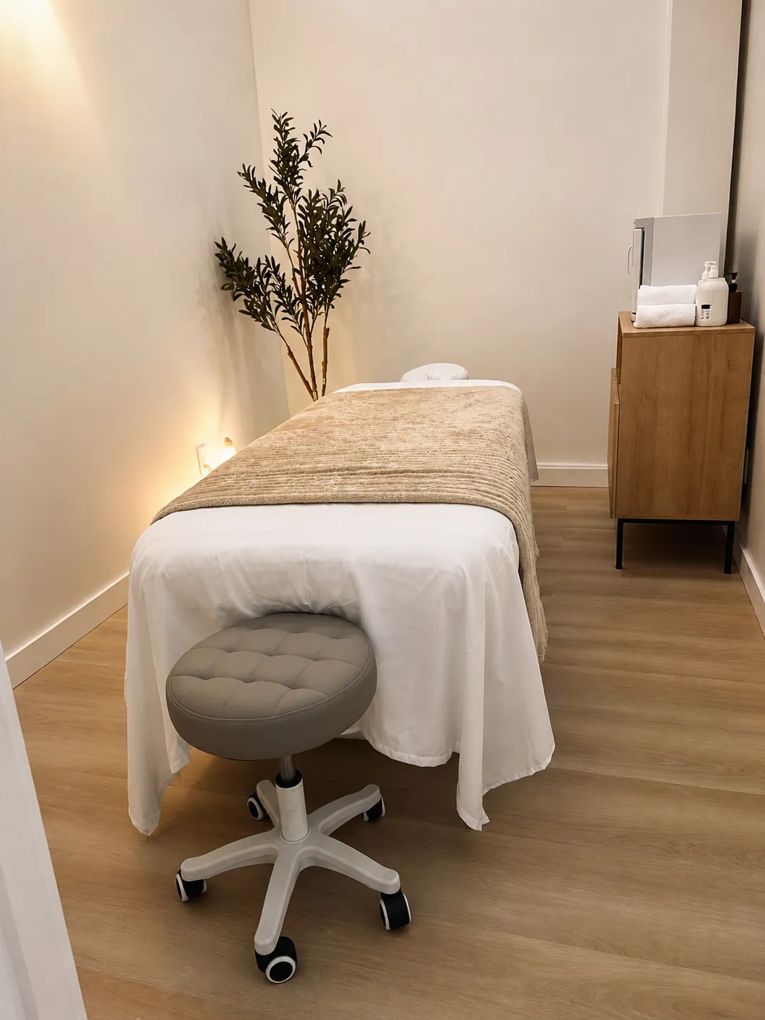 Treatment room at Harbor Wellness Massage in San Carlos with the table made up in white and tan, a wheeled stool and a warm towel cabinet